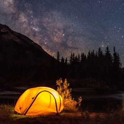 A tent glowing under a starry night sky, with a mountain and trees in the background.