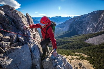 An ACMG Rock Climber High Above A Valley With Mountains In The Background