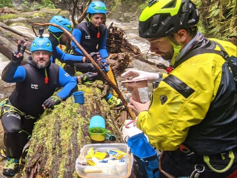 A Group Of People Wearing Canyoning Gear Eat Lunch Around A Log