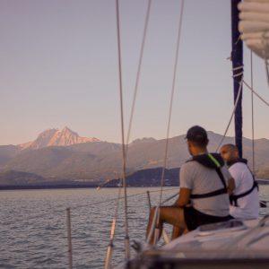 Two People Sat On A Sail Boat At Sunset With The Squamish Mountains And Howe Sound In The Background