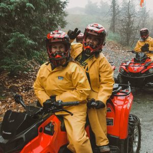 Two People Sitting On An ATV In Whistler BC, With Members Of Their Team Behind