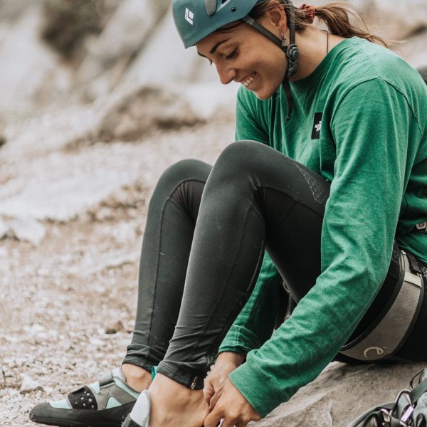 A woman wearing a climbing helmet is sat on the ground putting on rock climbing shoes with a smile on her face.
